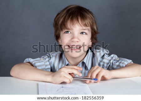 Happy schoolchild sitting at desk and writing in a notebook pen, portrait on gray background, studio, training