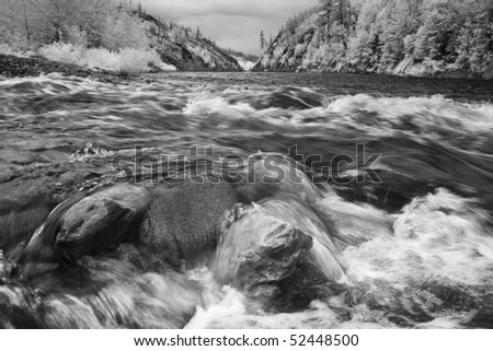 Black-and-white low angle view of river water rushing over rocks, with the tree-covered hills of Glacier National Park visible in the background. Horizontal shot.