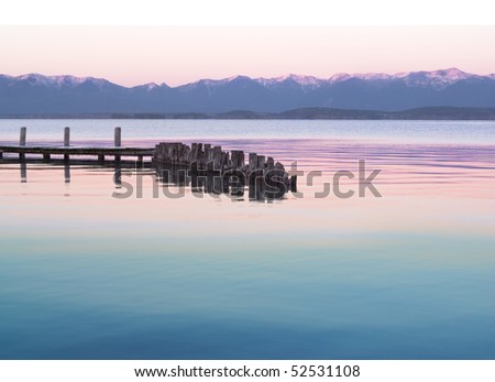 A wooden dock is extending out into a lake with calm water in the early morning. In the background can be seen hills and snow capped mountains. Horizontal shot.