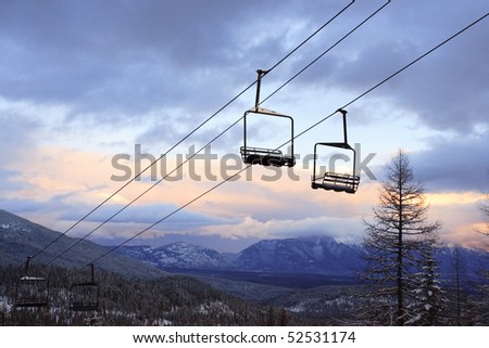 Empty chair lifts suspended above the ground over a ski slope. Trees and mountains can be seen in the background under a cloudy sky. Horizontal shot.
