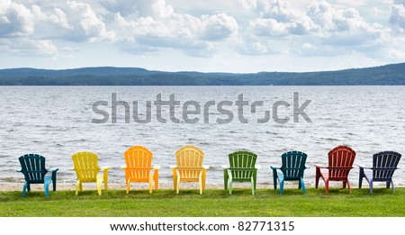 Eight colorful Adirondack chairs lined up on the beach looking out on the lake, mountains and clouds.