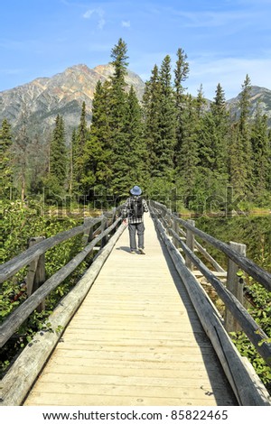 Man going on the wooden bridge through the river in a direction of wood and mountains