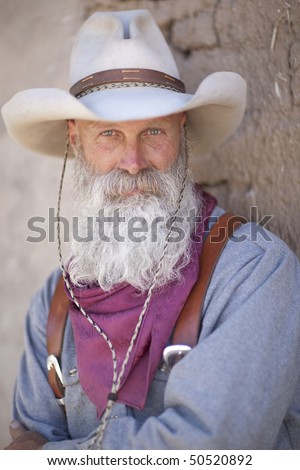 Portrait of a cowboy wearing a tall hat and sporting a long white beard. He is dressed in a heavy work shirt and kerchief. Vertical shot.