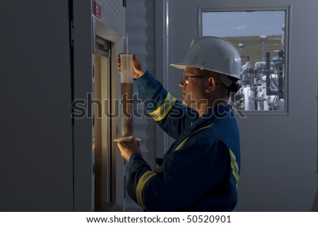 Petroleum worker measures a liquid in a graduated cylinder. He is wearing goggles and a hardhat. Horizontal shot.