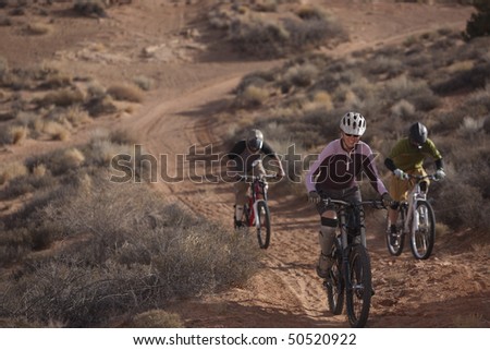 Three people are riding mountain bikes uphill in a desert landscape. Horizontal shot.