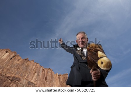 A smiling businessman rides a stick horse with one arm pointing to the sky in a desert landscape. Horizontal shot.