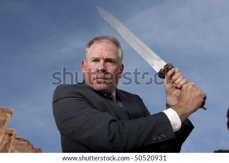 Businessman gives an intense look as he holds a sword, ready to battle. Horizontal shot.