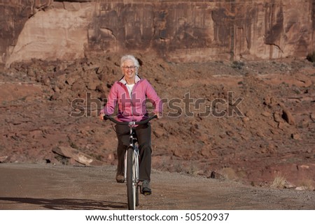 An elderly woman smiles as she rides her bike through a desert landscape. Horizontal shot.