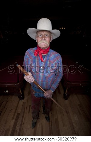 High angle portrait of a senior man in western wear, holding a pool cue and standing in a pool hall. He is looking at the camera with a serious expression. Vertical format.