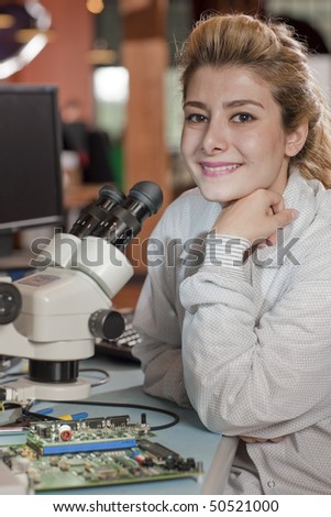 An attractive young researcher sits at a desk with a microscope used to look at electronics. She is smiling towards the camera. Vertical shot.