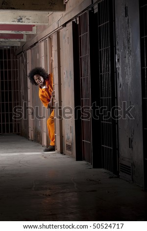 A young man wearing an orange prison jumpsuit makes a face as he peeks out from within his prison cell. Vertical shot.
