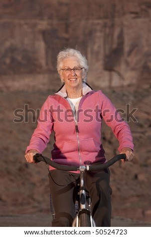 An elderly woman is sitting on a bike in a desert landscape and smiling at the camera. Vertical shot.