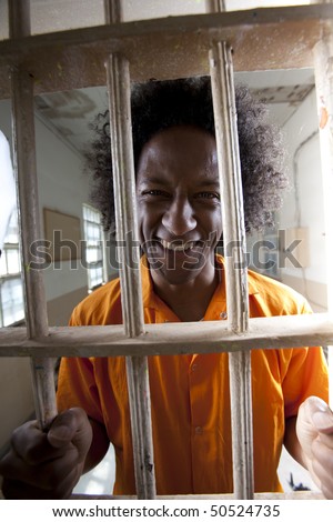 Portrait of a male prisoner with an orange jumpsuit and afro gripping the bars of a prison cell and smiling at the camera. Vertical format.