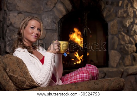 Portrait of a woman sitting in an armchair by a fireplace. She is holding a cup of tea and smiling at the camera. Horizontal format.