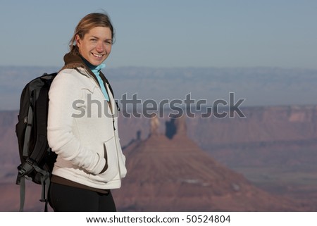 A young female hiker wearing a black backpack smiles at the camera.  In the background is a desert landscape. Horizontal shot.