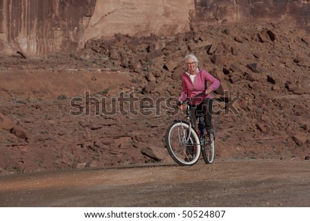 An elderly woman is sitting on a bike in a desert landscape and smiling at the camera. Horizontal shot.