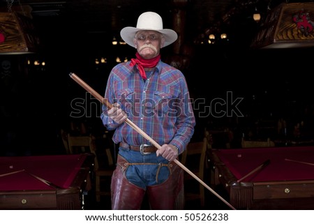 Portrait of a senior man in western wear, holding a pool cue and standing in a pool hall. He is looking at the camera with a serious expression. Horizontal format.