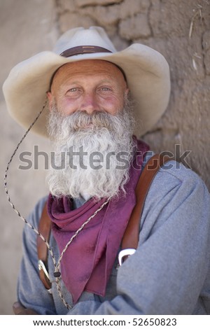 A senior man with a white beard is leaning against an adobe brick wall and looking at the camera. Vertical shot.