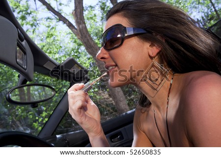 An attractive young woman in a convertible applying lipstick in the rear view mirror while driving. Horizontal shot.