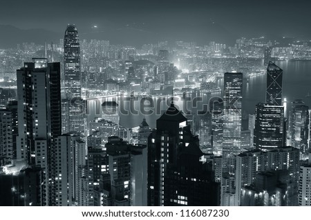 Hong Kong city skyline at night with Victoria Harbor and skyscrapers illuminated by lights over water viewed from mountain top in black and white.