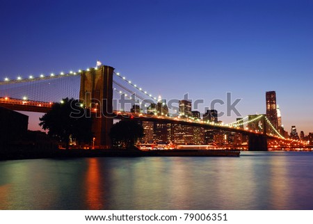 New York City Brooklyn Bridge and Manhattan skyline with skyscrapers over Hudson River illuminated with lights at dusk after sunset.