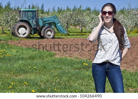 Young woman farmer speaking on her phone with a tractor in the field behind her.