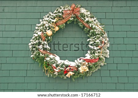 A snow covered Christmas wreath made of spruce and decorated and hung outside on the side of a building.