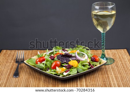 A plate of California salad containing oranges, dried cranberries, red onion, mixed lettuce, sliced almond and miniature tomatoes.  Beside it sits a glass of white wine.