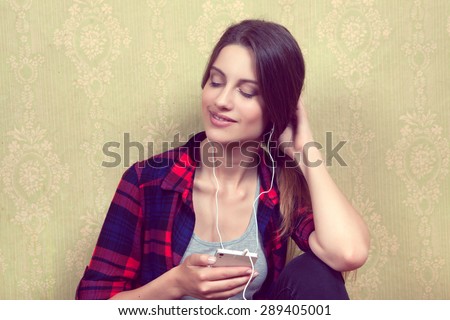 Mobile phone in the hands of a young girl with eyes closed wearing headphones and listening to music in a red plaid shirt sitting near the old yellow wall.