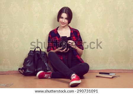 Old vintage film camera in the hands of a young girl in a red plaid shirt sitting near the old yellow wall.