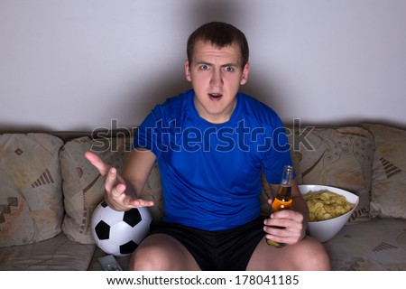 young shocked emotional supporter in uniform sitting on the sofa and watching football with beer and chips