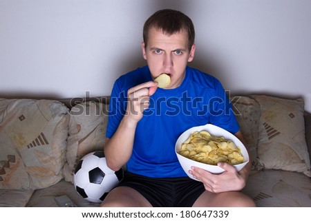 young supporter in uniform watching football on tv at home and eating chips