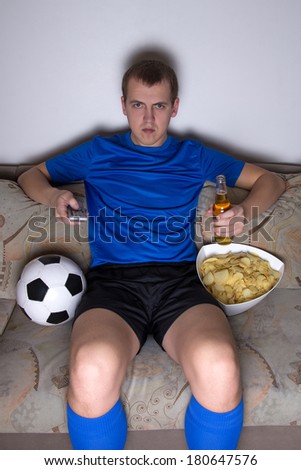 young bored man in football uniform sitting in living room and watching tv with chips and beer