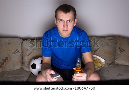 young football supporter in uniform sitting on the sofa and watching tv with beer and chips