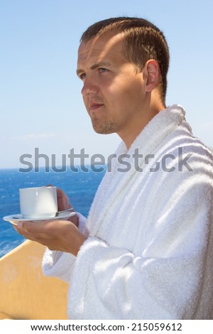 man in bathrobe with cup of coffee standing at terrace with sea view