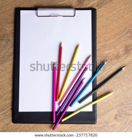 colored drawing pencils and clipboard with blank paper on wooden table background