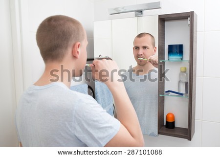 young man brushing teeth and looking at mirror in bathroom