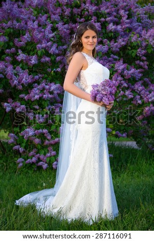 full length portrait of beautiful bride with long veil standing near blooming lilac tree in summer garden
