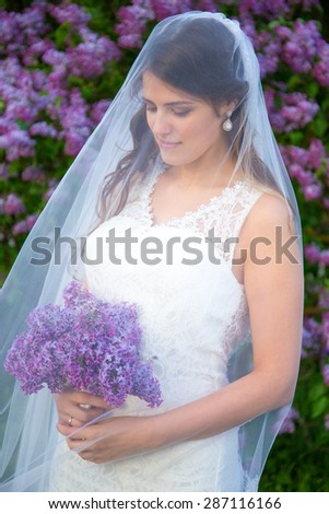young beautiful bride with long veil standing near blooming lilac tree