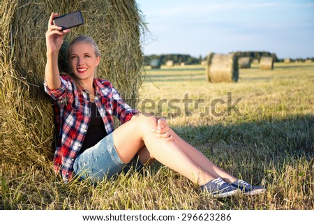 young beautiful blonde woman making selfie photo on smart phone in field with haystacks