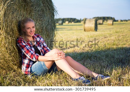 young beautiful blonde woman sitting near haystack in field
