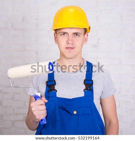 portrait of young man painter in work wear with roller paintbrush over white brick wall