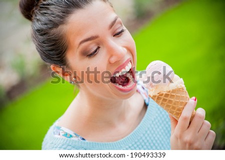 Attractive young brown haired happy excited girl in mint green sweater eating cold sweet ice cream in her free time on a bright summer day on green grass garden background