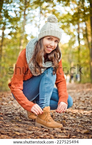 Young beautiful brown hiker girl in orange sweater and knitted hat tie the shoelace of her yellow boots in the cold autumn forest and enjoy the last warm hours