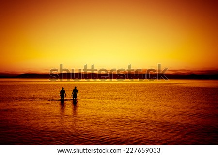 Two people walking away side by side in the beautifully rippled water refllecting the sky at  sunset, orange colored