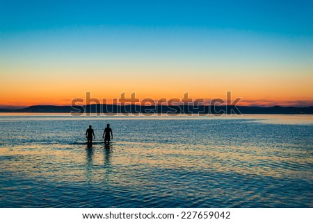 Two people walking away side by side in the beautifully rippled water refllecting the sky at  sunset