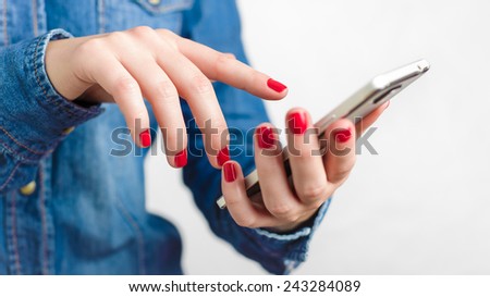 Young woman in blue denim shirt holding touch screen mobile phone in her red nail hands with small depth of field on homogenic blurred gray background