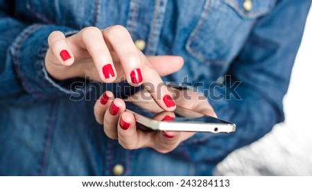 Young woman in blue denim shirt holding touch screen mobile phone in her red nail hands with small depth of field on homogenic blurred gray background