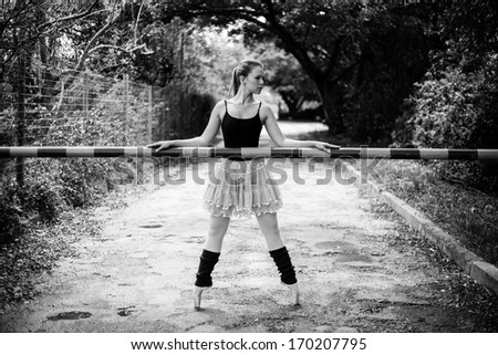 A beautiful young blond ballerina is standing on her toes in front of a boom gate like a security guard.This black and white photo has some beautiful classical lines.