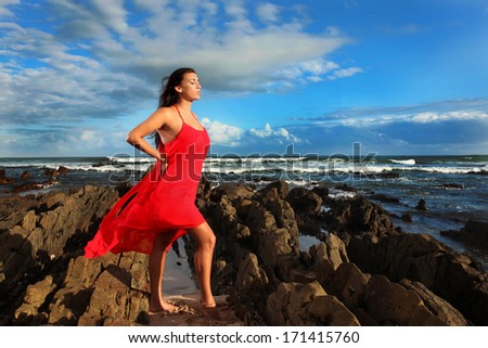 A beautiful brunette lady dressed in a long flowing red dress is soaking up the sun and the wind on a beach in Africa.